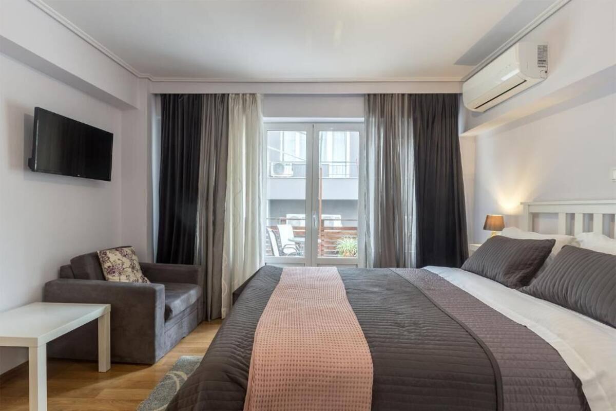 Quarto de hotel com cama, televisão, sofá e varanda com cortinas.