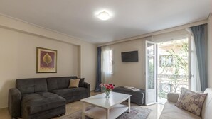 A living room with a grey sofa, a white coffee table, and a beige sofa. There is a framed picture on the wall, a TV mounted above the coffee table, and a balcony with a view of greenery. - UrbanStay (Athens)