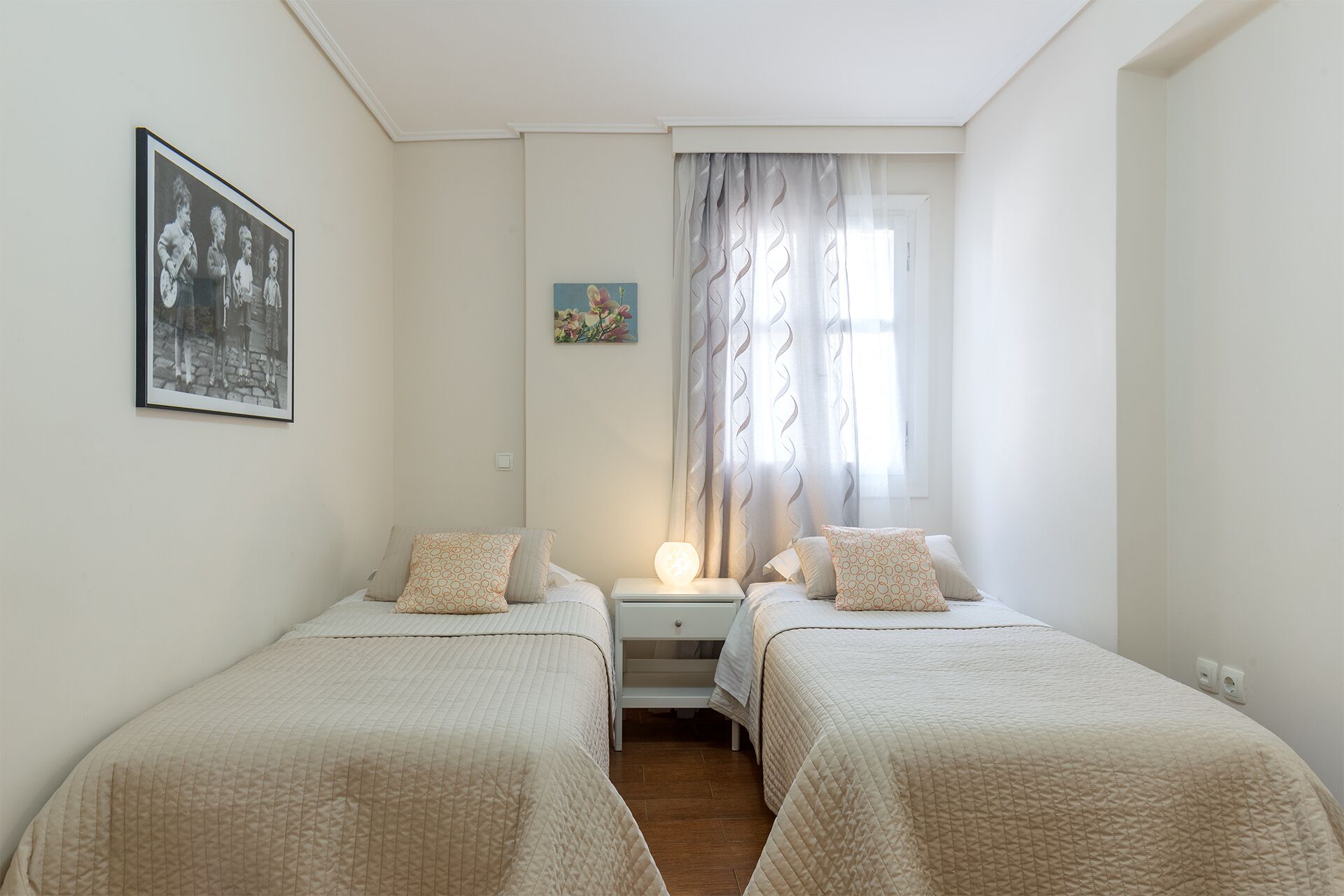 Un dormitorio con dos camas, una mesita de noche, una lámpara, una ventana con cortinas y un cuadro en la pared.
