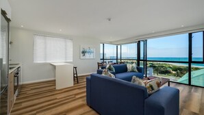 A modern living room with a large window overlooking a beach, a blue sofa, and a kitchen area with wooden flooring. - Paxton Luxury Apartments (Gqeberha (Port Elizabeth))