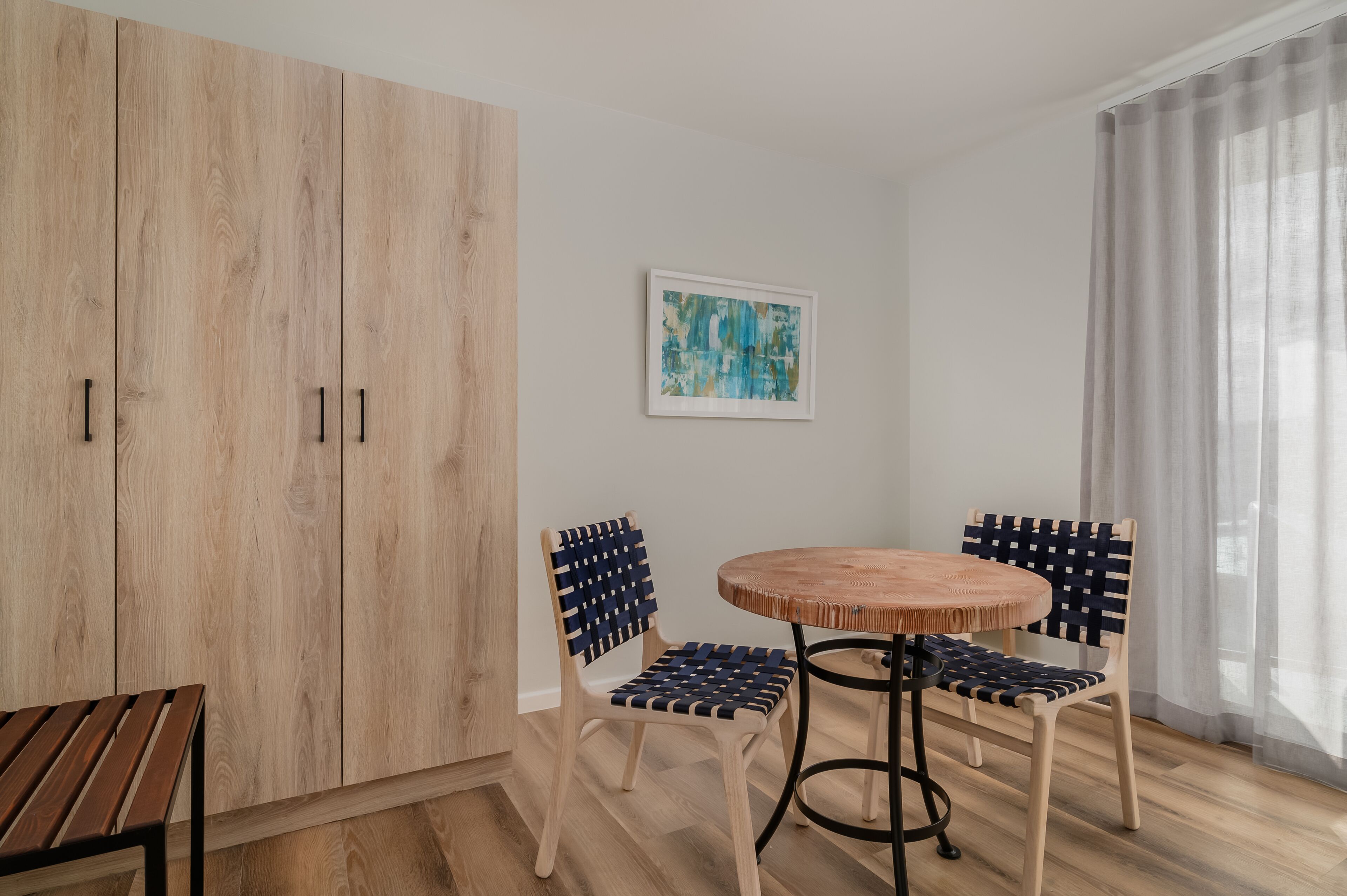 A room with a wooden wardrobe, two chairs with a woven seat, a round wooden table, and a framed abstract painting on the wall.