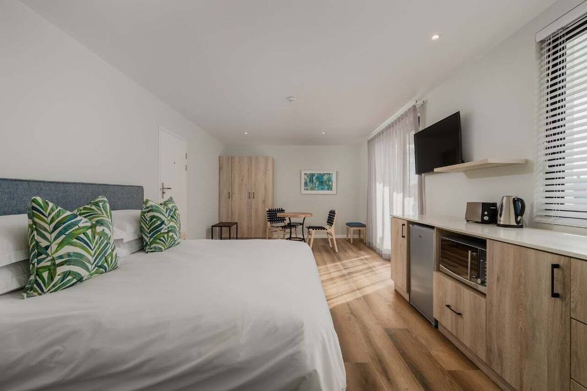 Ett modernt hotellrum med en säng, ett litet skrivbord, ett litet kök och en TV.