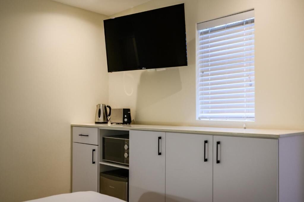A bedroom with a bed, a TV mounted on the wall, a window with blinds, and a kitchenette with a microwave and toaster.