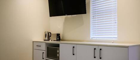 A bedroom with a bed, a TV mounted on the wall, a window with blinds, and a kitchenette with a microwave and toaster.