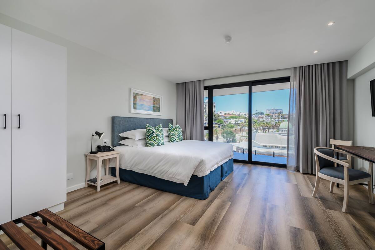 Ett modernt hotellrum med en stor säng, ett skrivbord och utsikt över staden genom ett stort fönster.