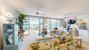 A spacious living room with a large sofa, a coffee table, and a TV. There is a balcony with a view of the water. - Evergreen 209 - Lower Level 3 Bdrm Lakefront Condo (Sunrise Beach)