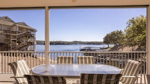 A balcony with a table and chairs, overlooking a body of water with a dock and a building in the background. - Evergreen 209 - Lower Level 3 Bdrm Lakefront Condo (Sunrise Beach)