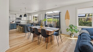 Eine moderne Küche mit weißen Schränken, ein Essbereich mit Holztisch und -stühlen und ein Surfbrett an der Wand.