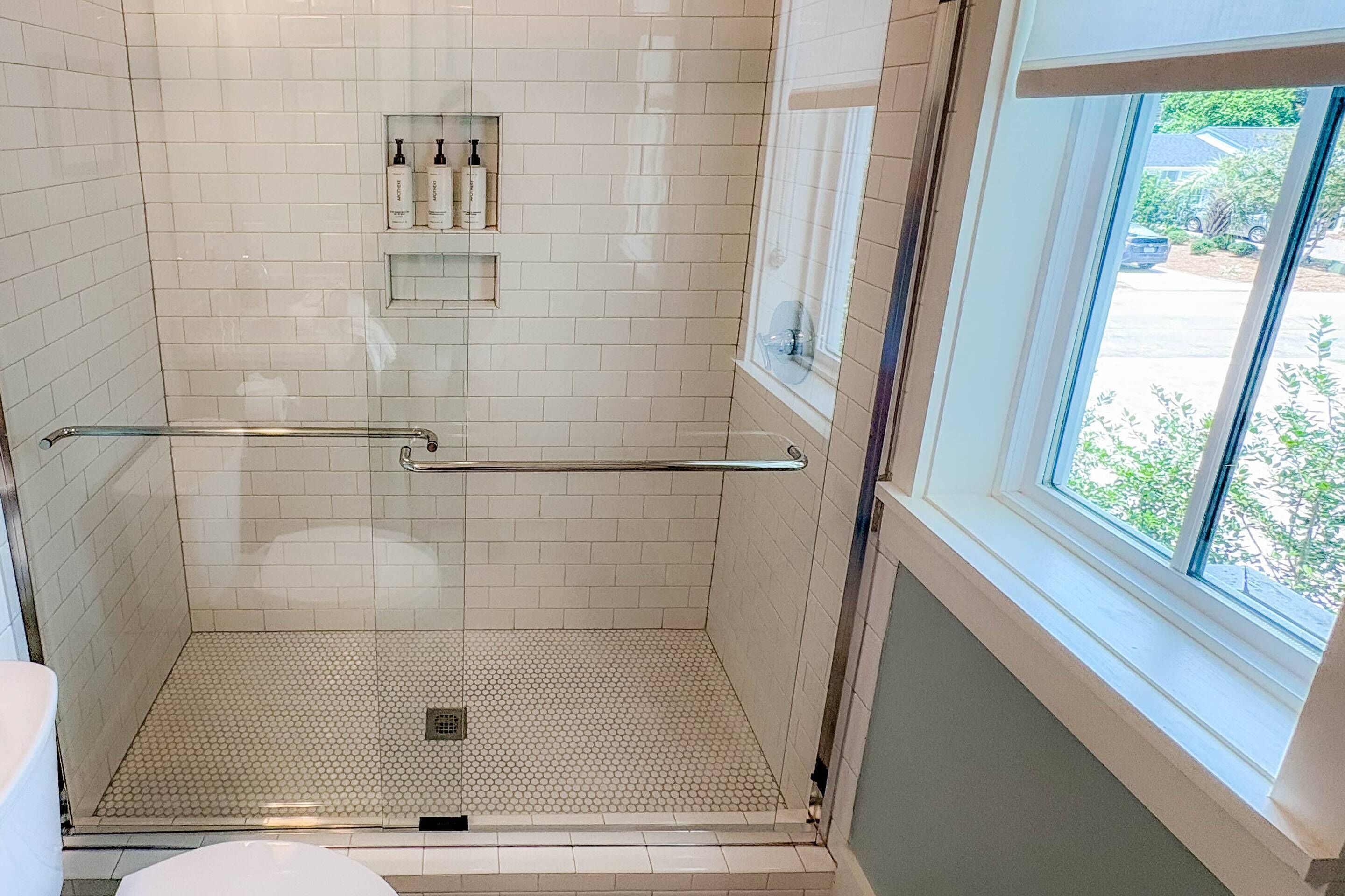 A shower with a glass door, a window, and a shelf with toiletries.