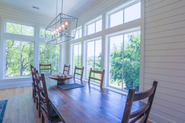 A dining area with a wooden table and chairs, large windows with a view of trees, and a hanging light fixture. - Private Pool & Hot Tub; Spacious Roof Deck w/ Views of Ocean;  2 Master Suites; 1 Block to Beach! (Isle of Palms)