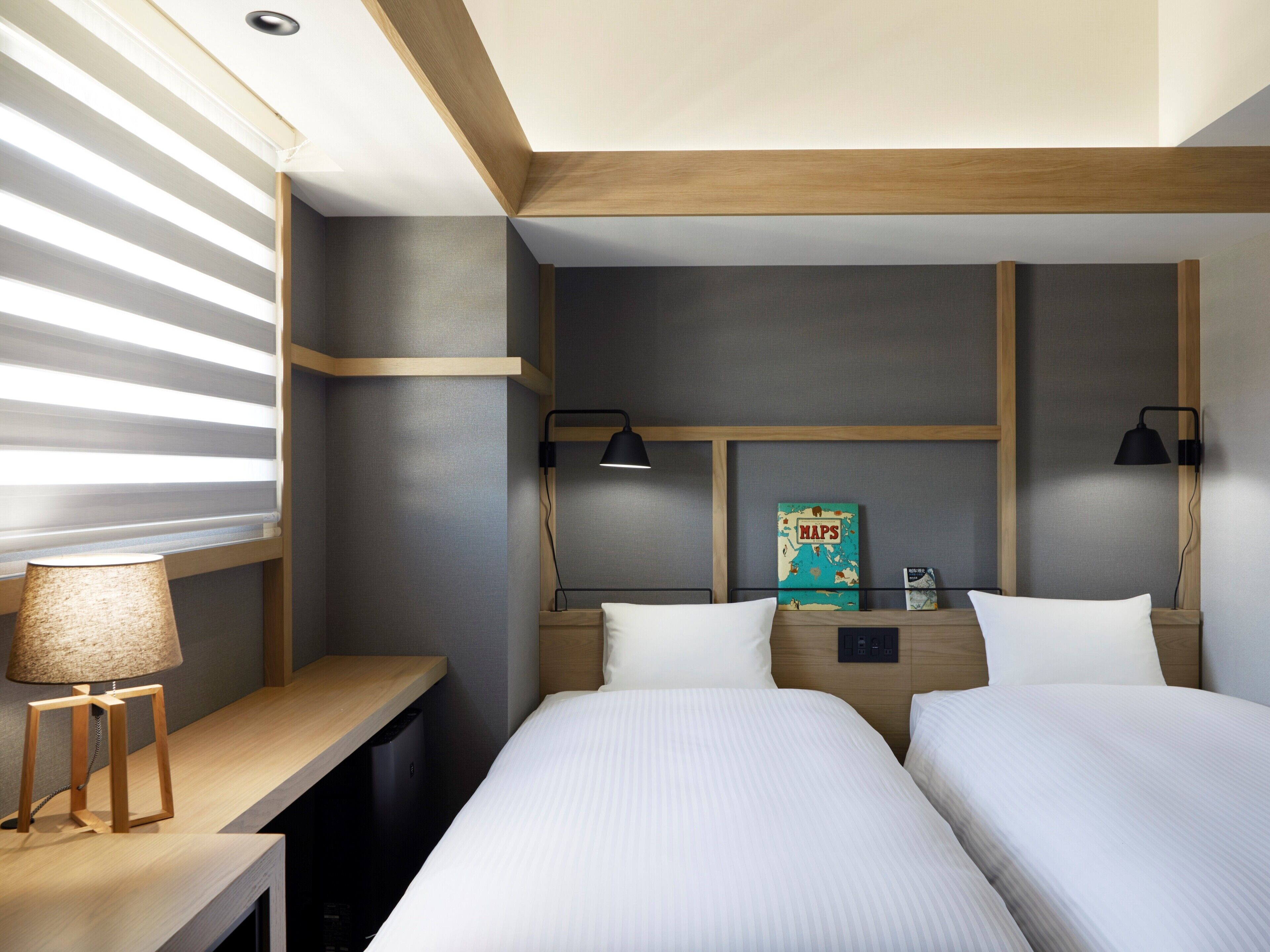 A hotel room with two beds, a desk, a lamp, a map on the wall, and a window with blinds.