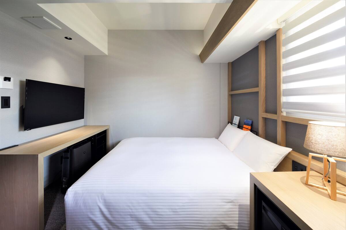 A modern hotel room with a large bed, a flat-screen TV mounted on the wall, a desk, and a window with blinds.