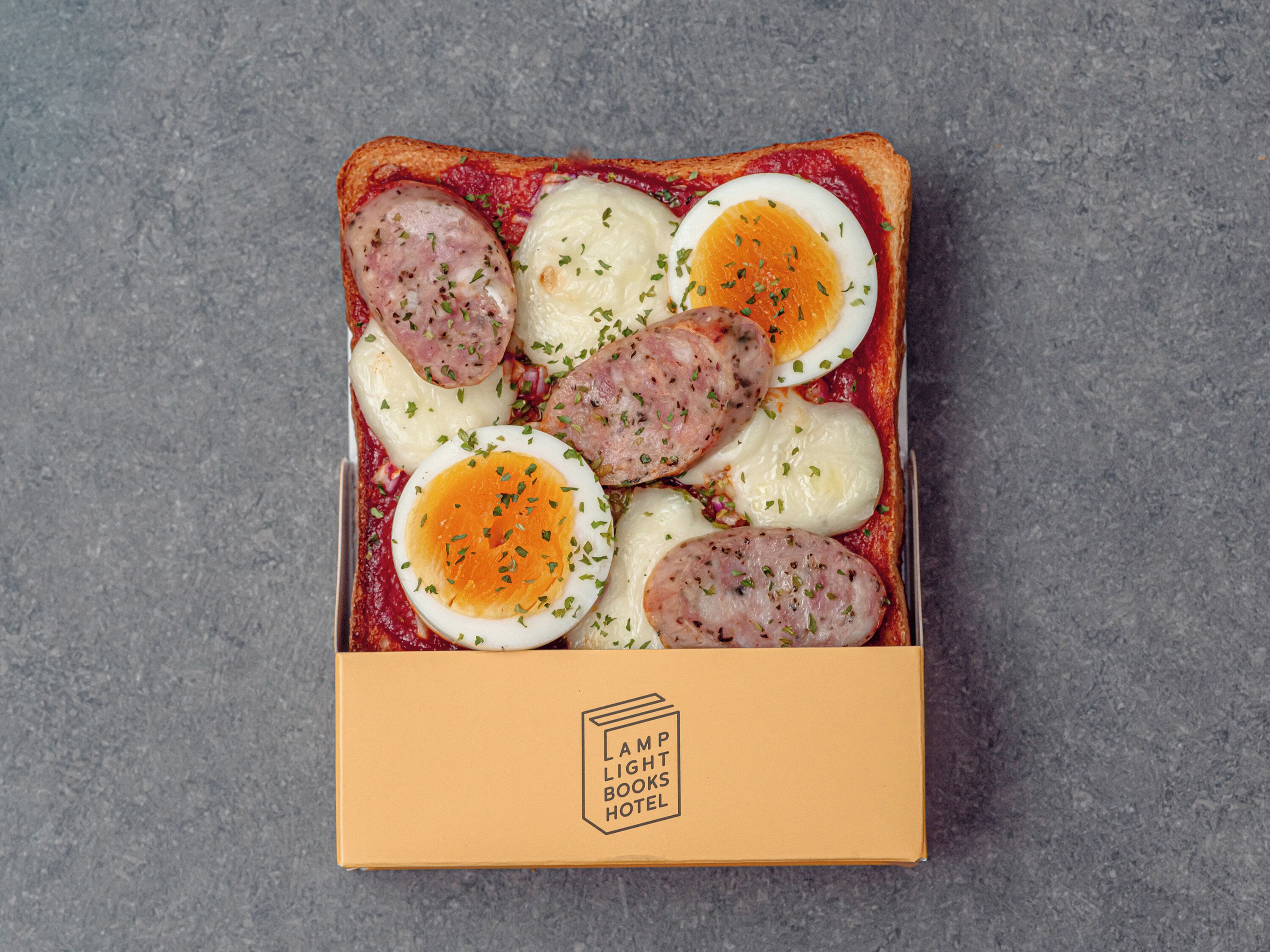 Una caja de comida con salchicha en rodajas, huevos cocidos y queso sobre una superficie rugosa.