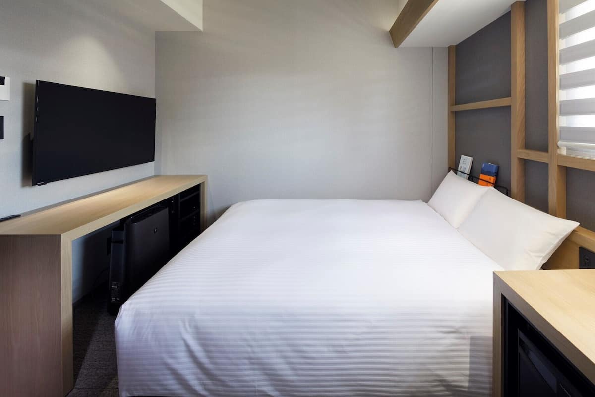 Habitación de hotel con cama, televisor montado en la pared, escritorio con lámpara y ventana con persianas.
