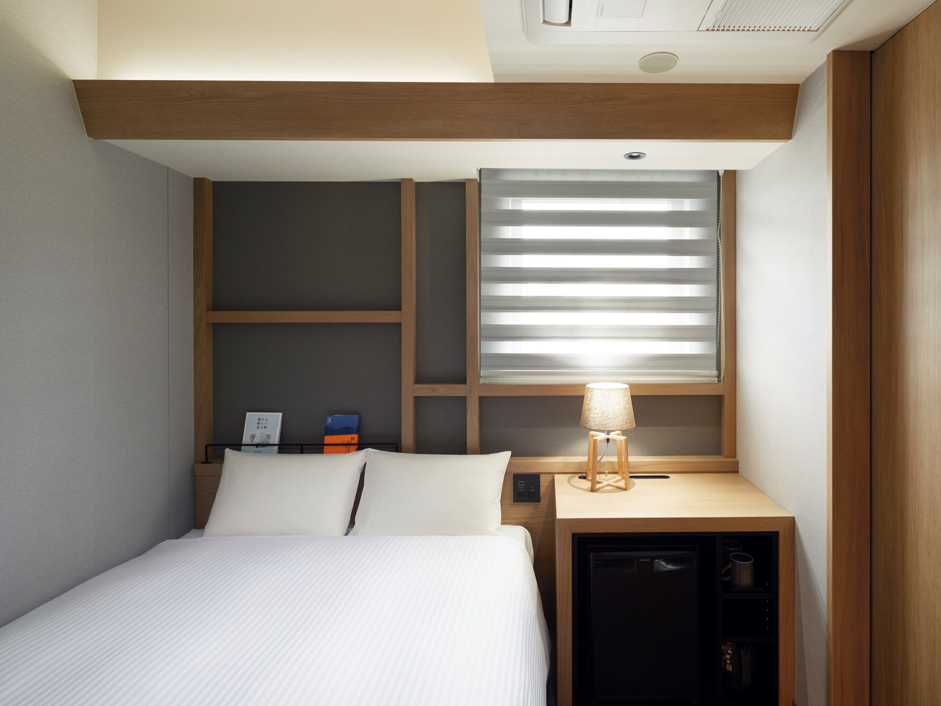 A compact hotel room with a bed, a small desk with a lamp, a built-in mini fridge, and a window with blinds.