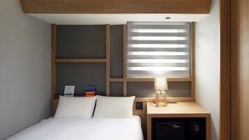 A compact hotel room with a bed, a small desk with a lamp, a built-in mini fridge, and a window with blinds.