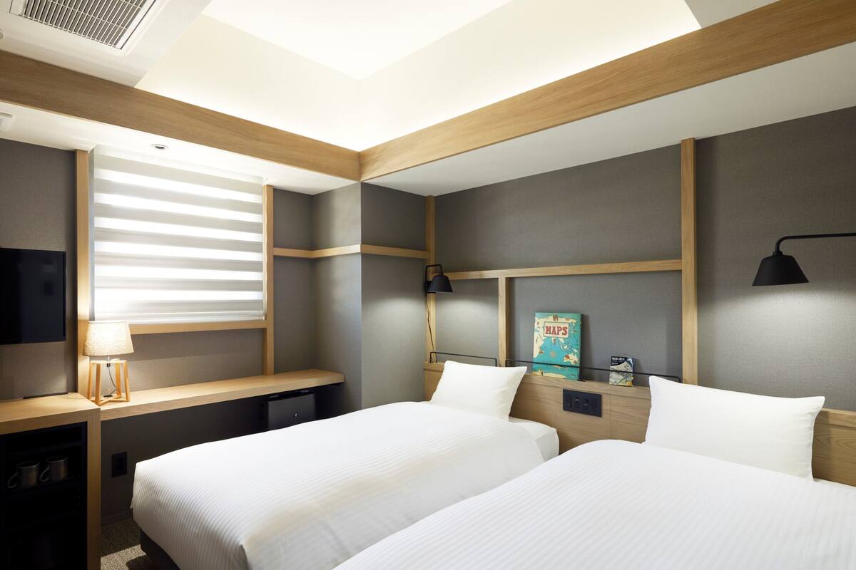 A hotel room with two beds, a desk, a TV, and a window with blinds.