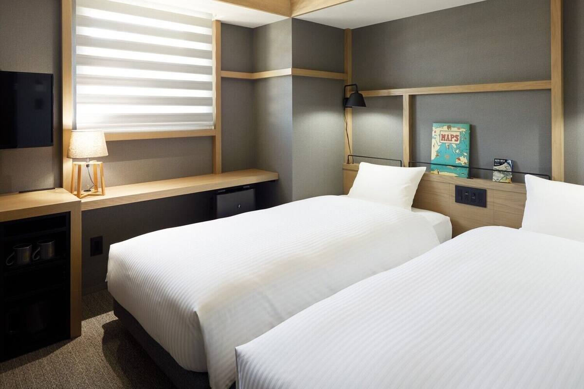 A hotel room with two beds, a desk, a lamp, a TV, and a map artwork on the wall.
