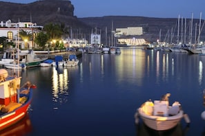 A marina with boats docked, buildings with lights, and a mountainous backdrop.