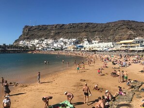 Ein überfüllter Strand mit badenden und sonnenden Personen, ein felsiger Hügel im Hintergrund und Gebäude entlang der Küste.