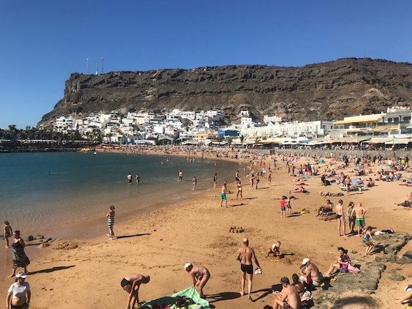 A crowded beach with people sunbathing and swimming, a rocky hill in the background, and buildings along the shore.