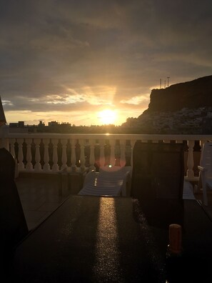 A balcony with a view of the sunset over a coastal town, featuring a railing and a table with chairs.