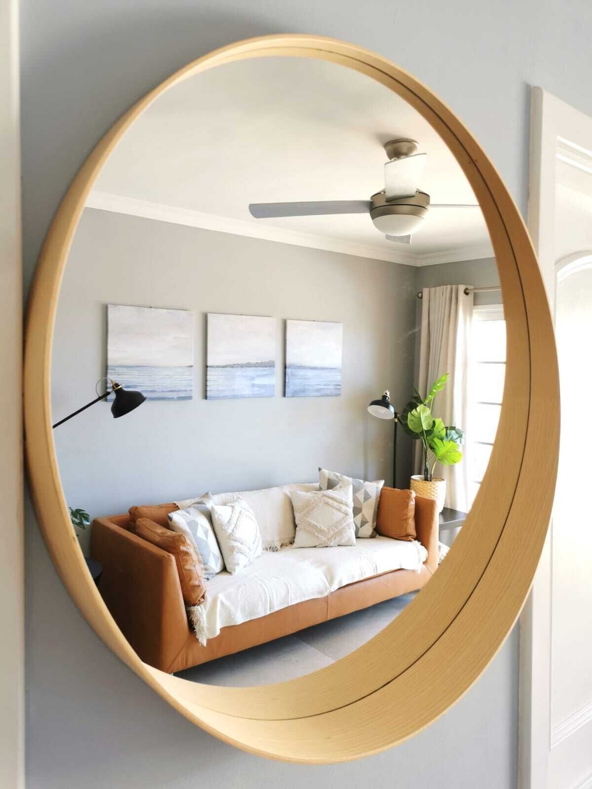 A room with a large oval mirror reflecting a light-colored sofa, a ceiling fan, and three wall-mounted artworks.
