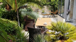 Un patio con una mesa de madera, sillas de mimbre y un paraguas rojo. Hay plantas en macetas y un muro de piedra al fondo.