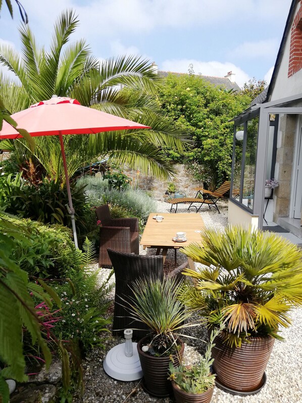 A patio area with a wooden table, wicker chairs, and a red umbrella. There are potted plants and a stone wall in the background.