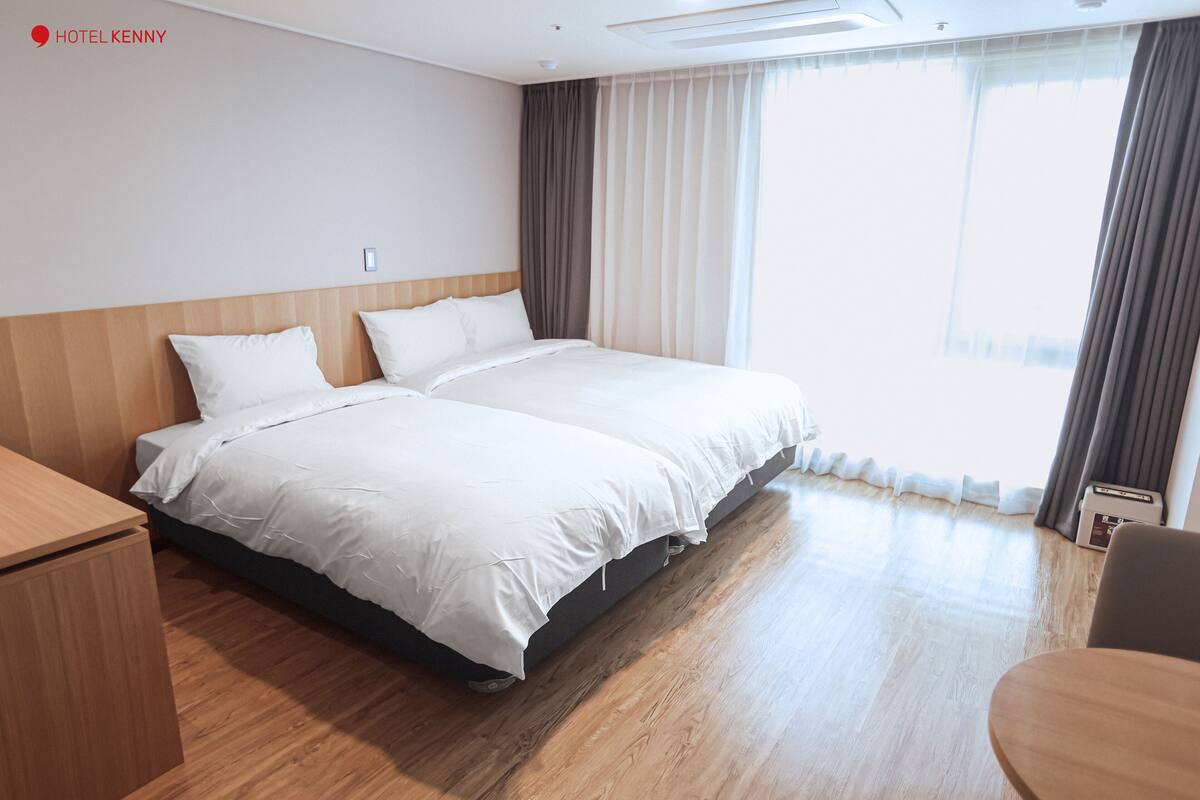 Quarto de hotel com cama de casal, cabeceira de madeira, mesa de cabeceira e uma janela grande com cortinas.