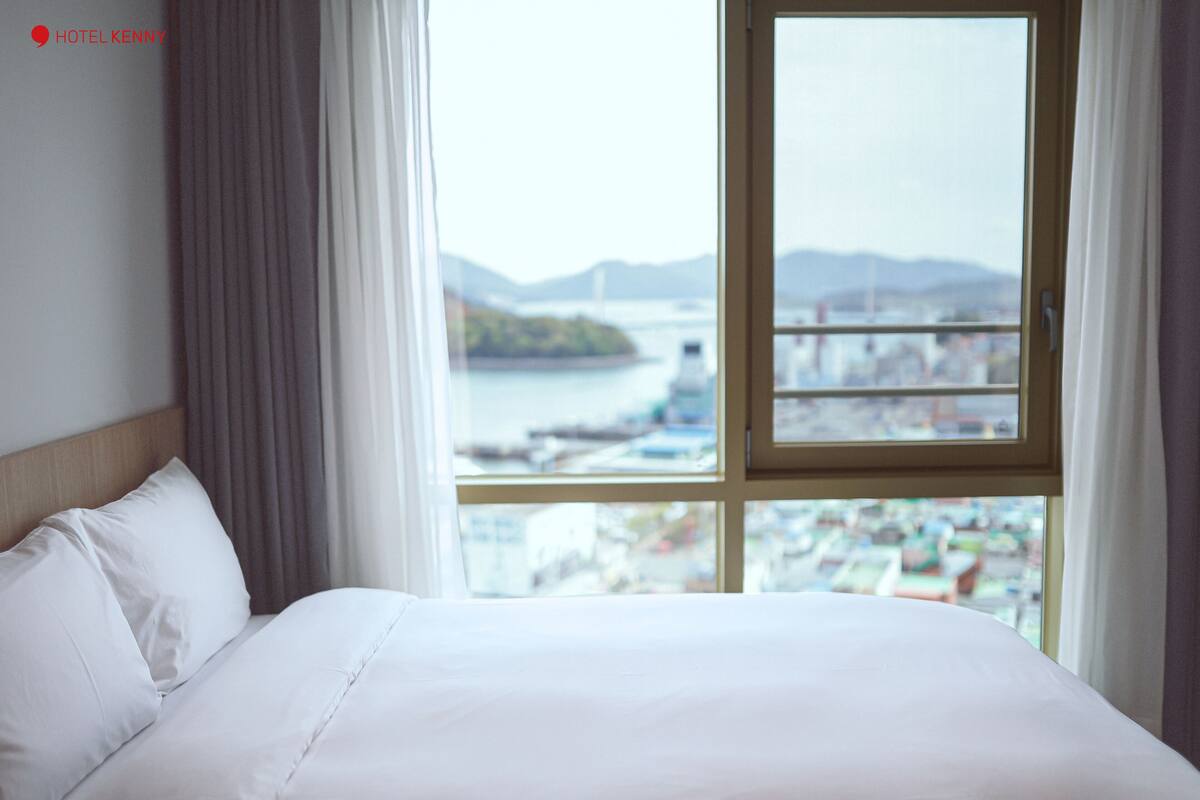 Quarto de hotel com uma cama grande, vista para a água e paisagem urbana ao longe.