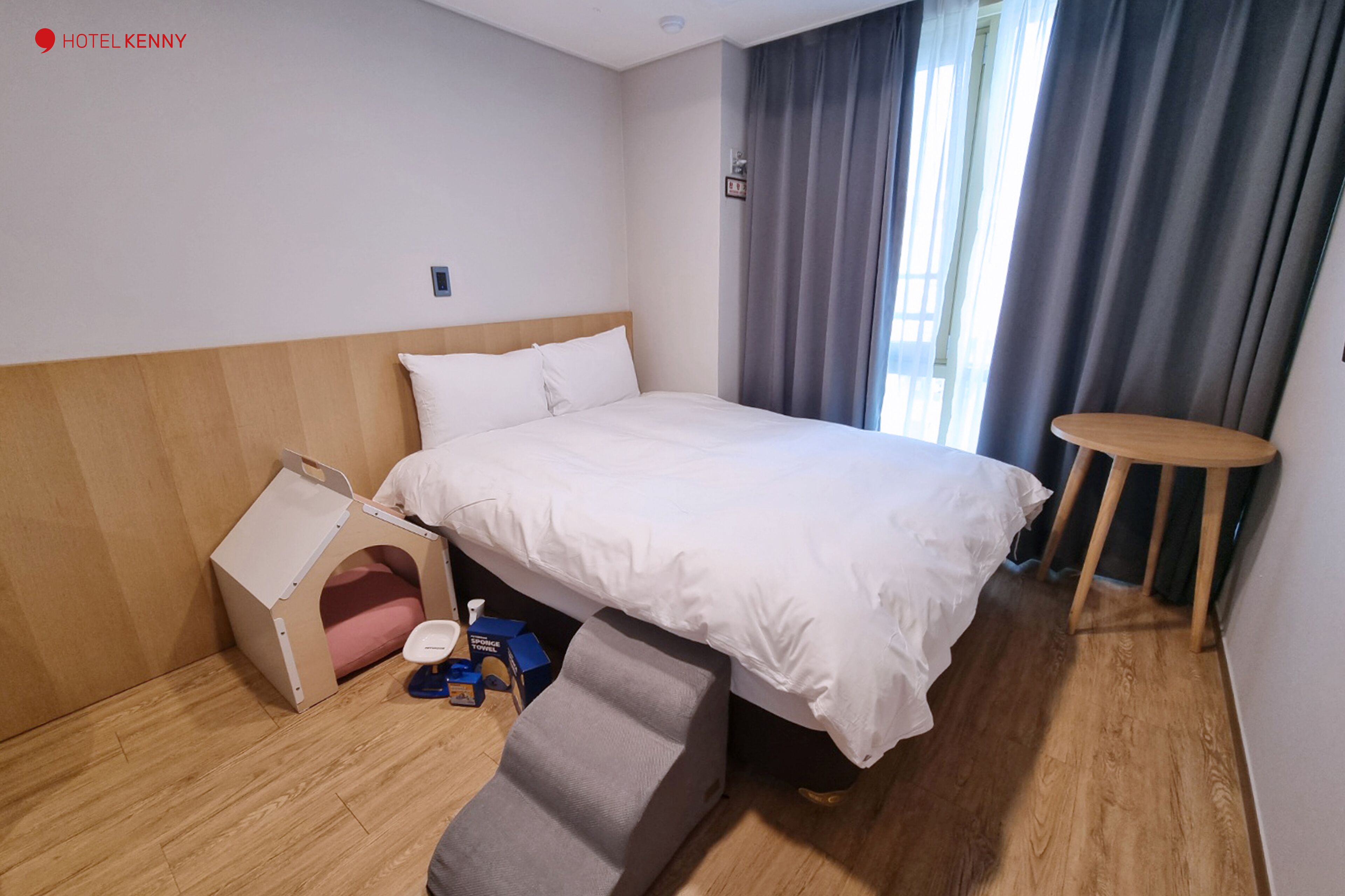 A hotel room with a bed, a small wooden side table, a footstool, a wooden headboard, and a window with curtains.