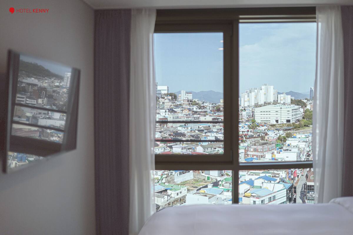 Quarto de hotel com uma janela ampla que oferece vista de uma área urbana densamente construída, com diversos edifícios e uma montanha ao longe.