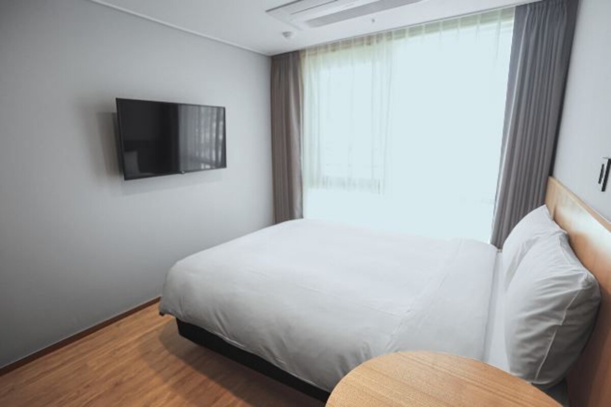 A hotel room with a bed, a TV mounted on the wall, a window with curtains, and a wooden floor.