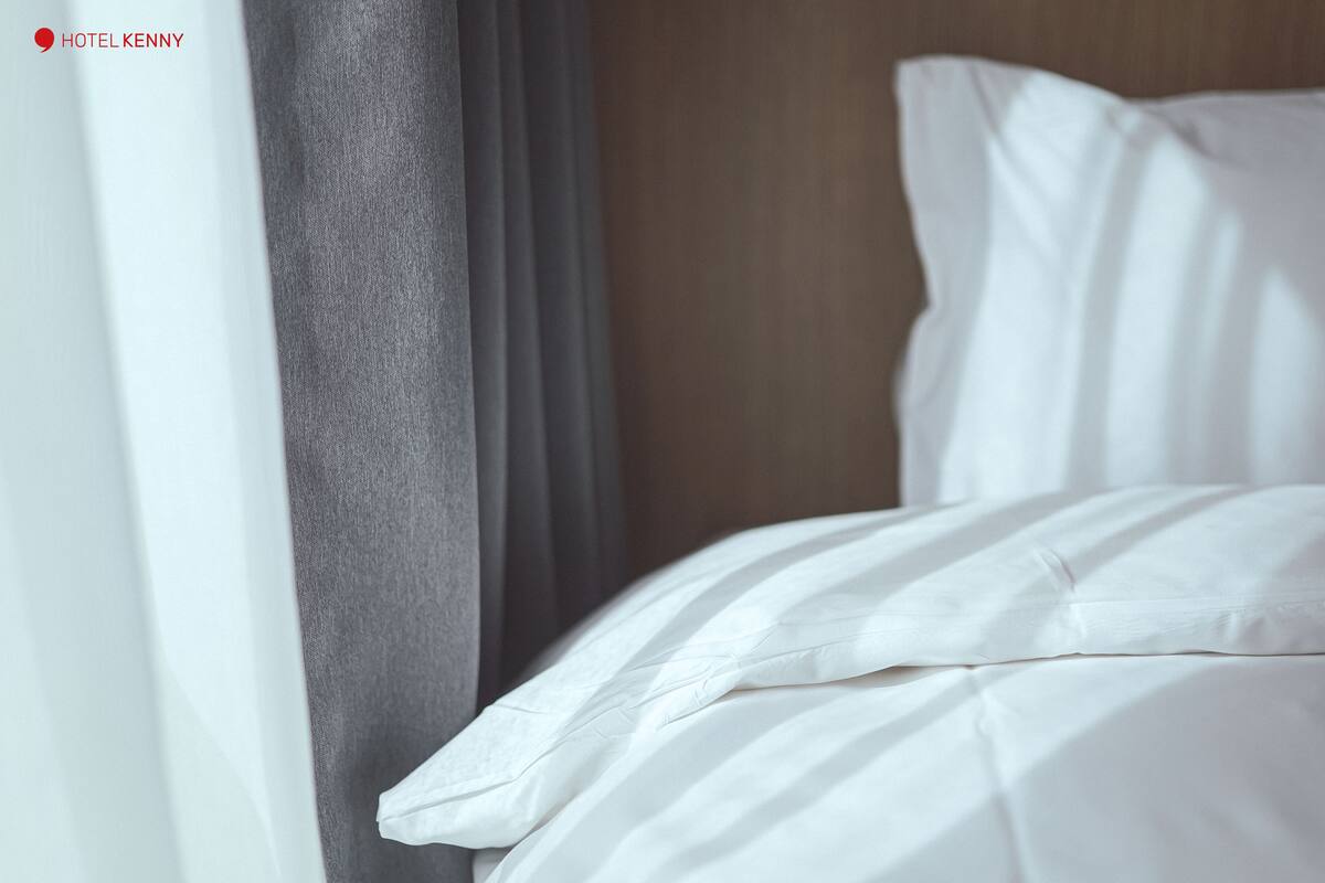 Uma cama bem arrumada com lençóis brancos, uma cortina cinza e uma cabeceira de madeira.