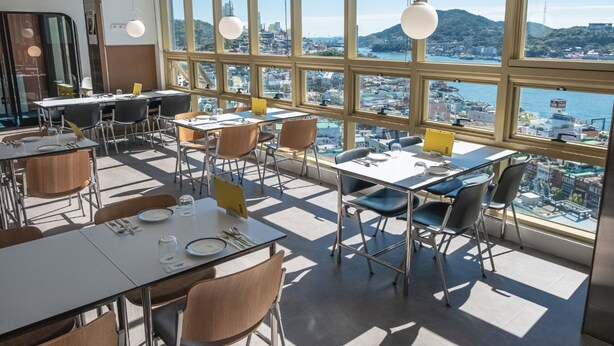A modern dining area with large windows offering a view of a coastal cityscape. - Hotel Kenny Yeosu (Yeosu)