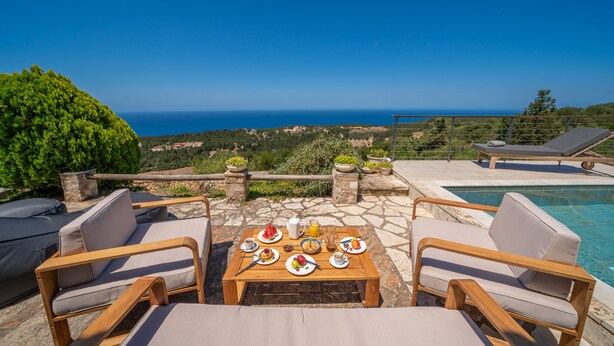 A wooden table with plates of food and drinks, surrounded by lounge chairs, overlooking a pool and a scenic view of the sea and greenery.
