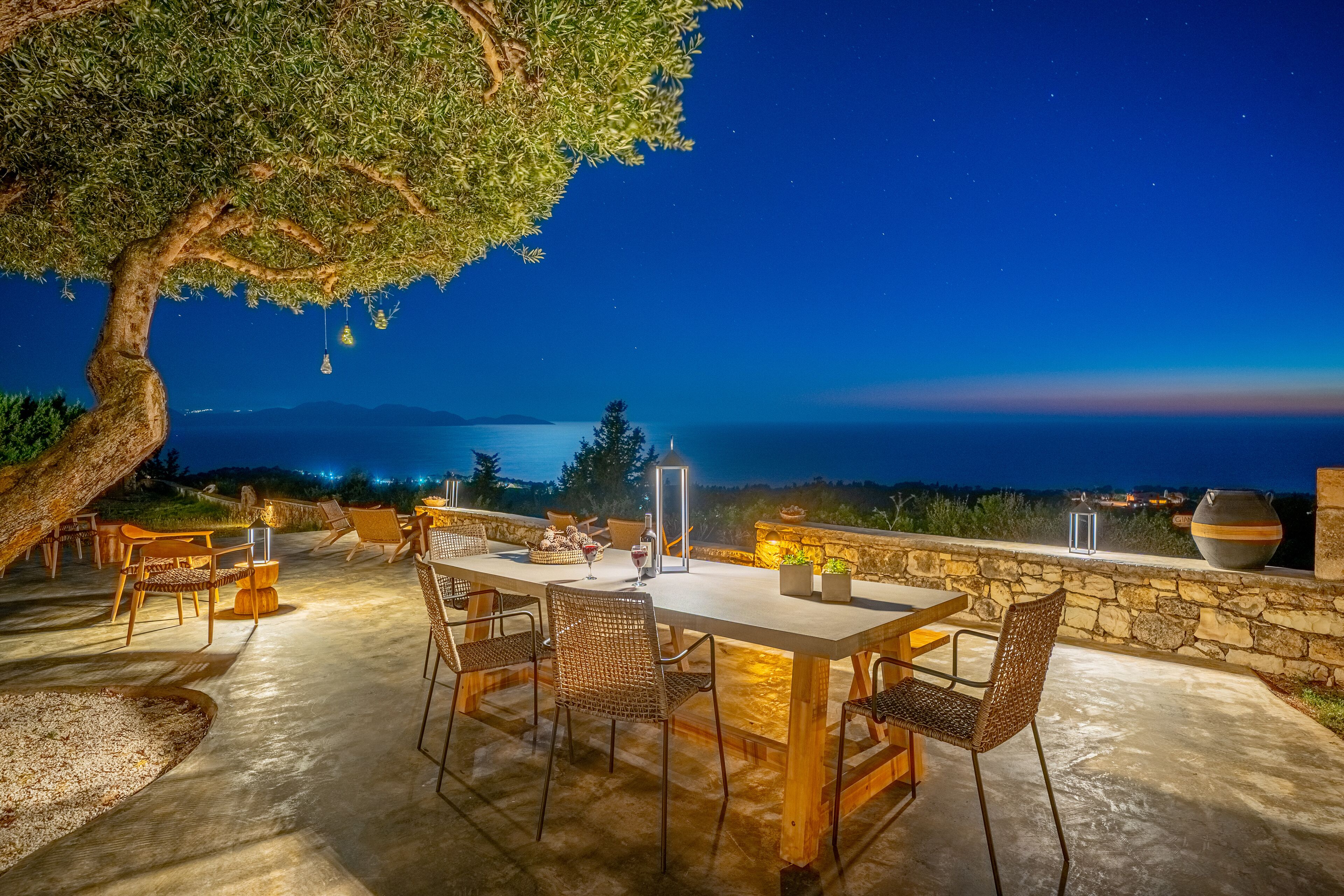 Un área de comedor al aire libre con una mesa y sillas, un muro de piedra y vistas al mar de noche.