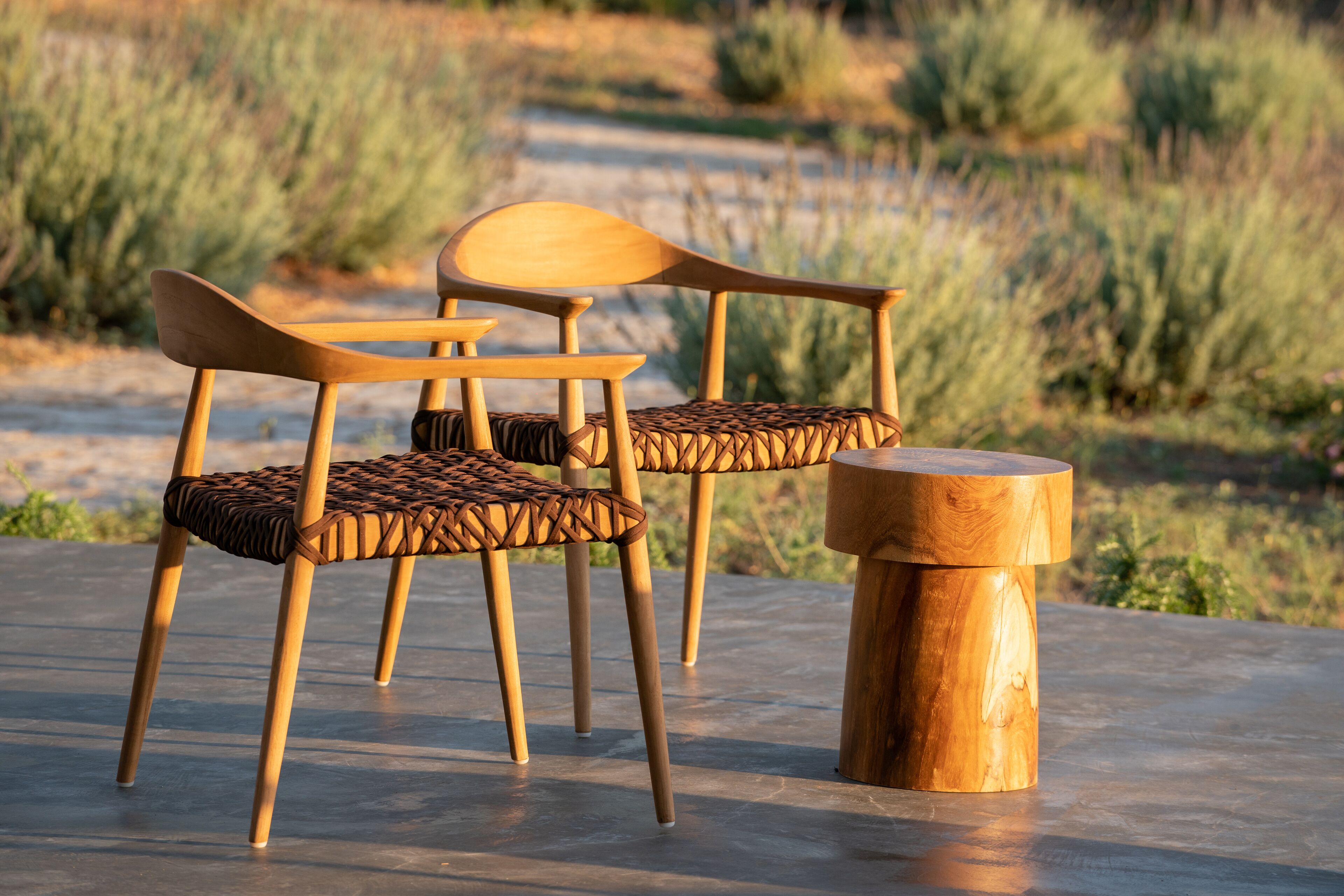 Two wooden chairs with woven seats and a round wooden table on a concrete surface.