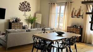 A modern living room with a sofa, dining table, and chairs. There is a staircase, a wall-mounted TV, and decorative items.