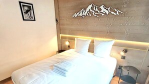 A neatly made bed with white linens and a folded towel, a wall-mounted light fixture, and a framed picture on the wall. - Flat for 4, central Les Gets, WiFi, parking (Les Gets)