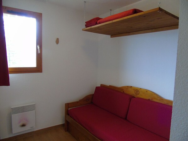 A small room with a wooden bed, a red sofa, a window, and a wall-mounted heater.
