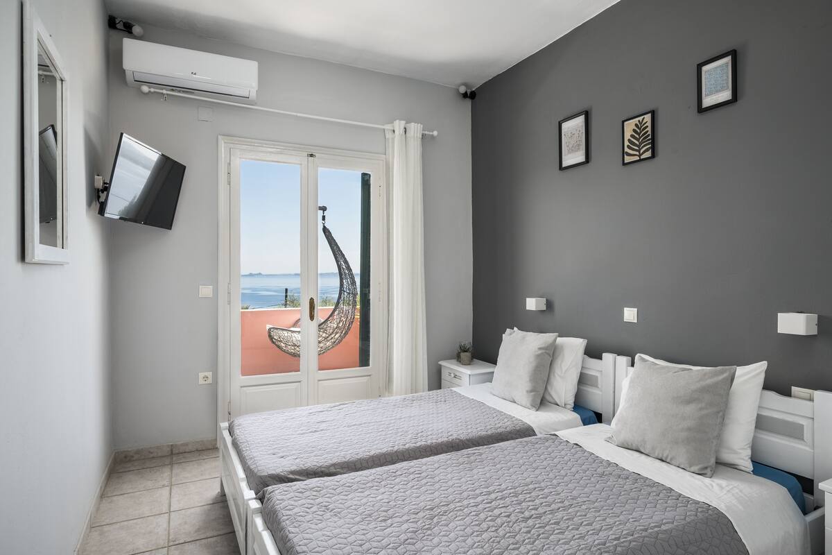 A bedroom with two beds, a TV mounted on the wall, a window with a view, and a ceiling fan.