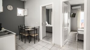 A modern kitchen with a dining table and chairs, a clock on the wall, and a view into a bedroom with a bed and a TV. - Ionian Sea View Apartments at Barbati by Konnect (Corfu)