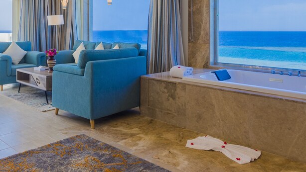 Una habitación de hotel moderna con un ventanal que ofrece vistas al océano, un sofá azul, una mesita con un jarrón de flores y una bañera con una toalla.