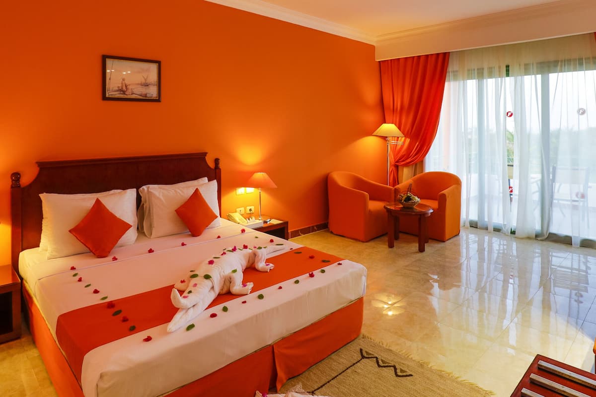 A hotel room with a large bed, two bedside tables, a nightstand, an orange armchair, a small table with a lamp, and a window with curtains.