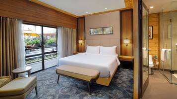 A hotel room with a large bed, a bench, a small table, a glass-enclosed shower, and a view of the outdoor area.