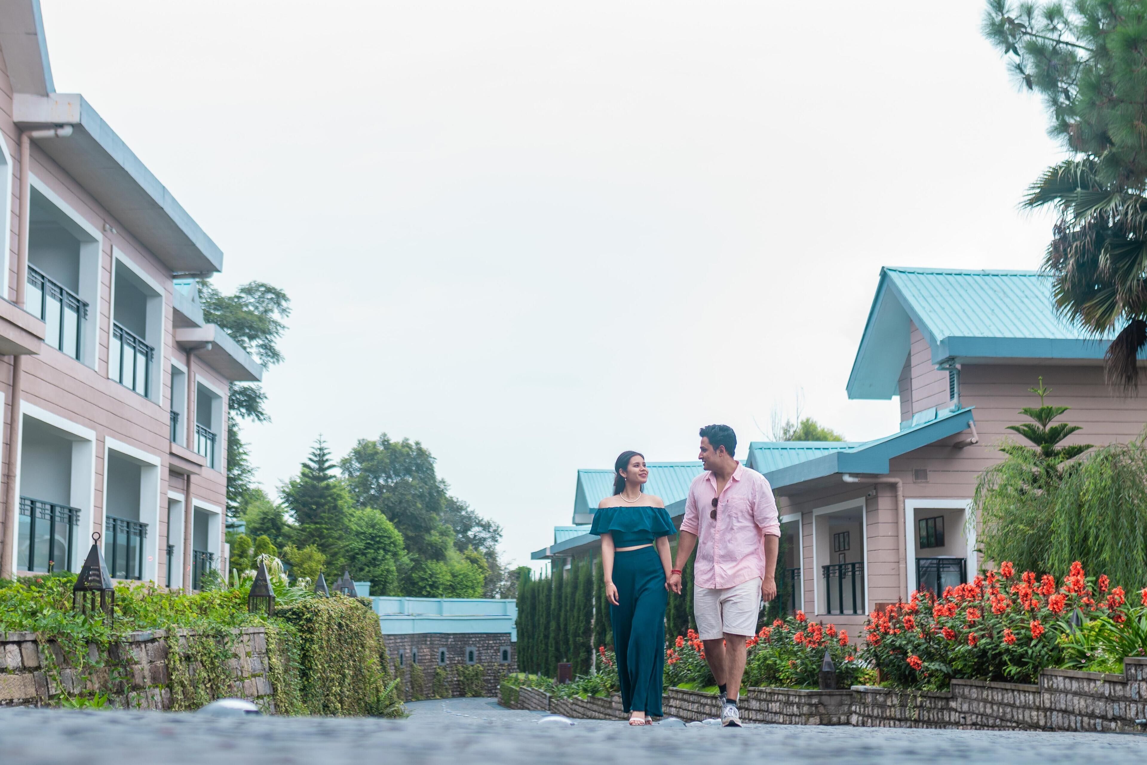 A couple walking on a pathway between buildings with balconies and a garden with red flowers.