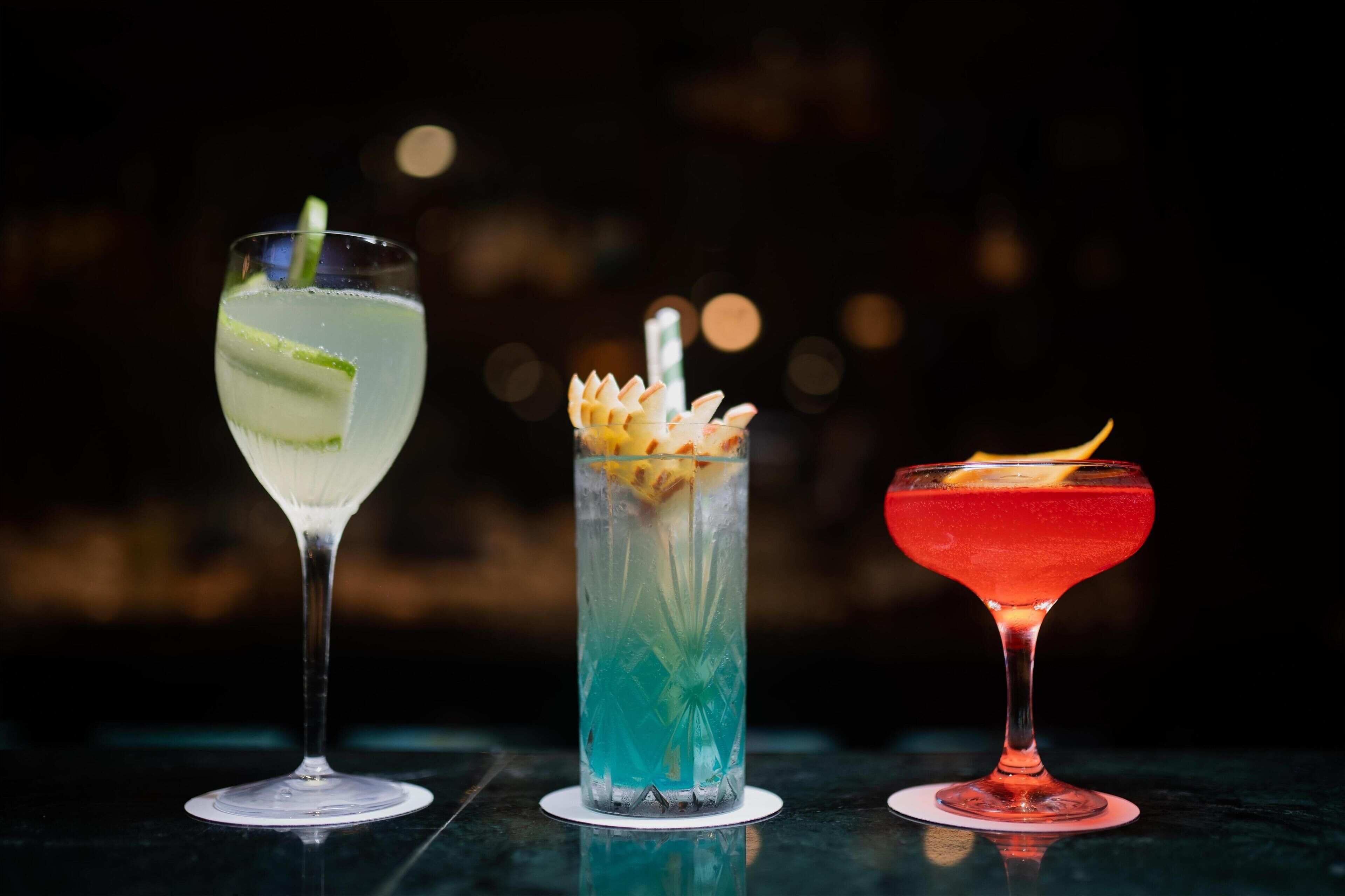 Drei verschiedene Cocktails in Gläsern auf einem Tisch vor dunklem Hintergrund.