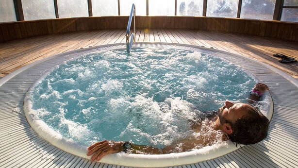 A man relaxing in a hot tub with a wooden deck and large windows overlooking a snowy mountain landscape.